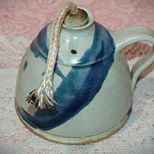 May include: A ceramic oil lamp with a rounded body, a side handle, and a thick braided wick extending from the top opening. The lamp features a light gray base color with bold, hand-painted dark blue brushstroke patterns wrapping around the exterior.