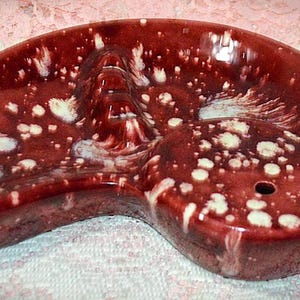 May include: A kidney-shaped ceramic ashtray with a glossy deep red glaze featuring white speckled patterns. The piece includes a central molded ridge for resting items and a single circular hole on the right side, finished with a smooth texture.