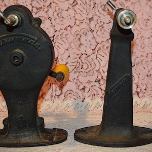May include: Two black cast iron Neumade film rewinding stands. The left unit features a circular housing with a hand crank and a yellow knob. The right unit is a vertical support arm. Both pieces have the Neumade brand name embossed on the metal.