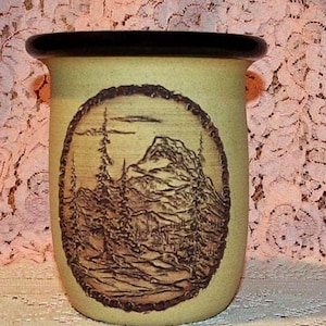 May include: A cylindrical ceramic vessel with a light tan body and a dark brown rim. The front features an oval-shaped embossed relief depicting a mountain landscape with tall pine trees and rocky terrain, rendered in a dark brown tone.