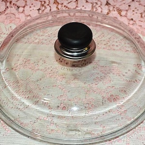May include: A round, clear glass lid for cookware featuring a centered black knob with a metallic silver base. The word PYREX is embossed in the center of the glass surface. The lid has a raised dome shape with a flat rim for a secure fit.
