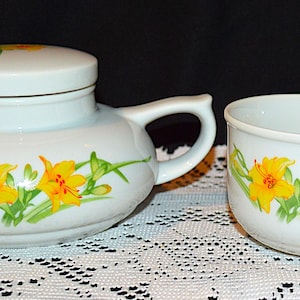 May include: A white teapot and teacup set with a floral design featuring yellow flowers and green leaves. The teapot has a lid and a handle. The teacup has a handle.