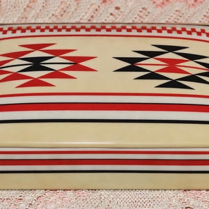 May include: A rectangular cream-colored box with a lid featuring a geometric pattern in red and black. The design includes two large diamond-shaped motifs on the top, bordered by horizontal stripes and a small checkered band along the edge.