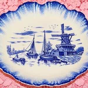May include: An oval ceramic platter with a scalloped edge featuring a blue and white transferware pattern. The central illustration depicts a coastal scene with sailboats, a large windmill, and small buildings, all rendered in deep blue ink.