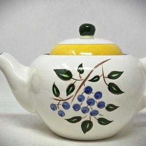 May include: A white ceramic teapot featuring a hand-painted design of blue berries and green leaves on the side. The teapot has a curved handle and spout, and it is topped with a bright yellow lid that includes a small, dark green knob handle.