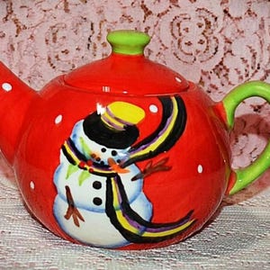 May include: A bright red ceramic teapot featuring a hand-painted snowman wearing a black hat and a colorful striped scarf. The teapot has a lime green handle, a matching green lid knob, and white polka dots scattered across the red surface.