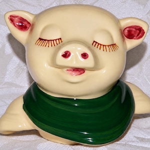 May include: A cream-colored ceramic pig figurine featuring a glossy finish, closed eyes with painted brown eyelashes, a snout with red nostrils, a small red mouth, and a dark green scarf wrapped around its neck area, presented in a front view.
