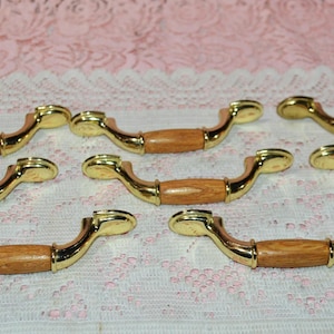 May include: Eight cabinet pulls with a gold finish and wooden center sections. The pulls have a curved design and are arranged on a pink and white patterned surface. The hardware is likely for furniture.