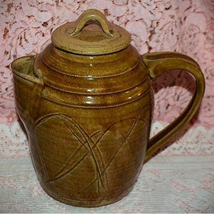 May include: A brown glazed ceramic teapot featuring a cylindrical body with a wide handle and a matching lid. The exterior surface is decorated with incised, overlapping curved lines that create a subtle pattern across the front of the vessel.