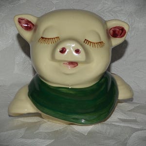 May include: A cream-colored ceramic pig figurine featuring a glossy finish, closed eyes with painted brown eyelashes, a pink snout, and a dark green scarf wrapped around its neck. The pig has small ears with reddish-brown accents on the inner parts.