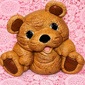 May include: A ceramic bear figurine with a textured light brown finish, featuring dark brown accents on the ears and feet. The bear has large, glossy black eyes, a black nose, and an open mouth showing a pink tongue, depicted in a seated position.
