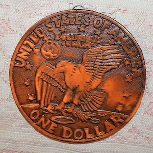 May include: A copper-colored, circular wall hanging featuring a detailed relief of a United States one-dollar coin. The design includes an eagle with outstretched wings, the words "UNITED STATES OF AMERICA," and "ONE DOLLAR."