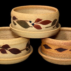 May include: Three round ceramic bowls with matching saucers, each featuring a textured beige finish and a hand-painted horizontal band of dark red and brown leaves. The bowls have a thick, rounded rim and a consistent cylindrical shape.