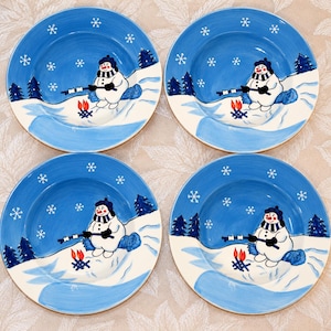 May include: A set of four round ceramic plates featuring a blue winter sky with white snowflakes. Each plate depicts a snowman wearing a blue hat and scarf, roasting marshmallows over a small campfire near dark blue pine trees on snowy ground.