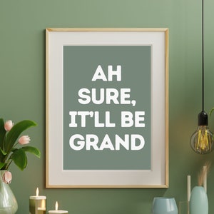 以下が含まれることがあります： セージグリーンの背景に白い文字で「Ah sure, it'll be grand」と書かれた額装されたプリント。プリントは、セージグリーンの壁に、キャンドルや花瓶のある木製のキャビネットの上にかけられています。