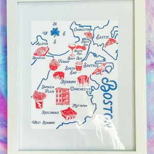 May include: Framed map art of Boston, Massachusetts, with blue and red illustrations of landmarks and food items. The map includes neighborhoods like Allston, Brighton, and Roxbury, with the city name in a stylized font.