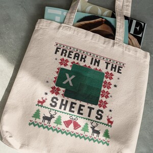 Puede incluir: Bolso tote beige con un diseño festivo. La bolsa presenta el texto "FREAK IN THE SHEETS" con un logotipo de Excel, rodeado de gráficos temáticos navideños como renos, campanas y copos de nieve. Se ven revistas dentro de la bolsa.