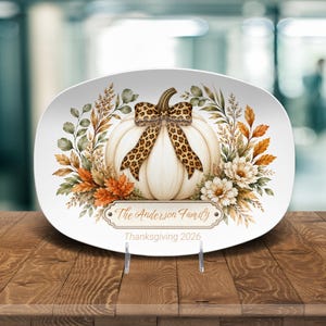 May include: An oval white serving platter featuring a central cream-colored pumpkin adorned with a leopard print bow. The design includes autumn foliage, flowers, and a decorative plaque with the text The Anderson Family and Thanksgiving 2026.