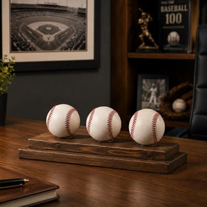 May include: Three white baseballs with red stitching are displayed in a row on a rectangular, dark brown wooden stand. The stand consists of two stacked wooden tiers, providing a stable base for the three baseballs to sit upright.