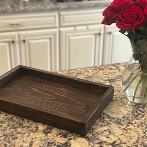 May include: A rectangular, dark stained wooden tray with a raised edge. The tray is empty and sits on a granite countertop. A glass vase with red roses is in the background.
