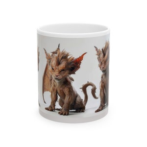 May include: White ceramic mug with a colorful illustration of a mythical creature with brown fur, orange wings, and a long tail. The creature has a fierce expression and is looking directly at the viewer.