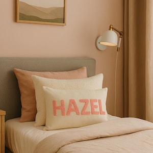 Puede incluir: Interior de dormitorio con una cama con ropa de cama y almohadas de color rosa claro. Un cojín rectangular crema con el nombre "HAZEL" en letras color coral está colocado sobre la cama. También se ven un cuadro de paisaje enmarcado y una lámpara de pared blanca.