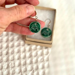 Pode incluir: Um par de brincos de cerâmica verde com um design de montanha e sol. Os brincos estão sobre um fundo branco.