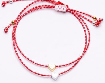 May include: A double-strand bracelet made of red and white twisted cord featuring one small gold-toned heart charm and one small silver-toned heart charm. The bracelet includes an adjustable sliding knot closure with small metallic beads at the ends.