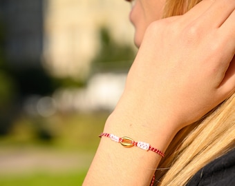 May include: A bracelet featuring a central gold-colored cowrie shell charm flanked by small white woven segments, attached to a thin red and white braided cord. The cord wraps around the wrist, showcasing a simple and delicate design.
