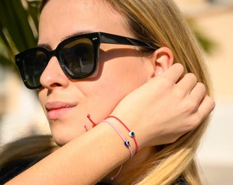 May include: Two thin cord bracelets worn on the wrist, one in bright red and one in light pink. Each bracelet features a small, centered evil eye charm with a blue, white, and black circular design. The cords are adjustable with loose ends.