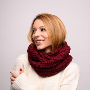 Puede incluir: Una mujer con un suéter blanco y una bufanda infinita de color granate. La bufanda está hecha a crochet y tiene un aspecto texturizado.