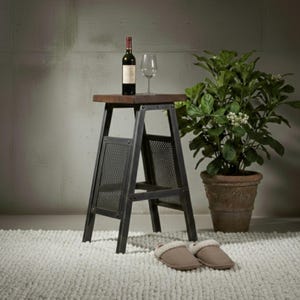 Op de afbeelding: Een kleine bijzettafel in industriële stijl met een donkerhouten blad en een zwart metalen frame. Een fles wijn en een wijnglas staan op de tafel. Een potplant en pantoffels staan in de buurt. De tafel is ongeveer 61 cm hoog.