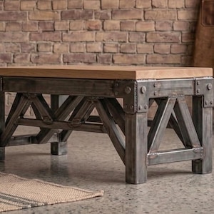 Peut inclure: Table basse de style industriel avec un plateau rectangulaire en chêne et une base en acier rivetée. La base présente un design géométrique avec des supports inclinés. La table est placée contre un mur de briques.