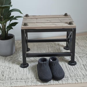 Könnte beinhalten: Ein quadratischer Beistelltisch mit einer Holzplatte und einem schwarzen Metallrahmen. Die Oberseite ist mit großen Schrauben befestigt. Ein Paar graue Strickhausschuhe liegt auf dem Teppich unter dem Tisch. Der Tisch ist etwa 46 cm hoch.