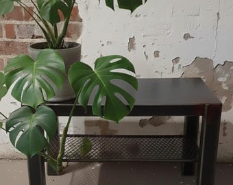 Könnte beinhalten: Ein dunkler Metall-Beistelltisch mit einem unteren Regal, auf dem eine Topf-Monstera-Pflanze mit großem, grünem, geschlitztem Blattwerk steht. Der Tisch steht vor einer Ziegel- und weißen Wand.