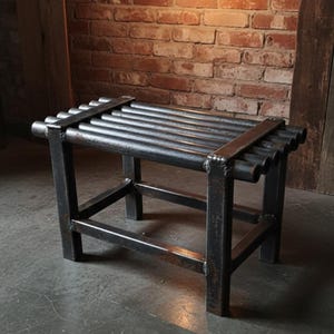 Può includere: Panca in stile industriale in metallo scuro. La seduta è composta da tubi metallici paralleli, sostenuti da un robusto telaio. La panca è posta contro un muro di mattoni. Misura circa 90 cm di lunghezza.