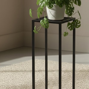 Peut inclure: Un support de plante en métal noir avec un dessus et une base carrés, soutenant un pot blanc texturé rempli d'une plante verte et feuillue. Le support est sur un tapis beige, créant une esthétique moderne et minimaliste.