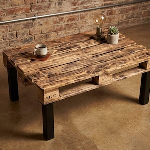 Könnte beinhalten: Ein rechteckiger Couchtisch aus einer recycelten Holzpalette mit dunkler, geflammter Oberfläche. Der Tisch verfügt über vier stabile, mattschwarze Metallbeine und zeigt eine sichtbare Holzmaserung sowie eingeprägte Markierungen.