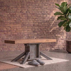 Op de afbeelding: Een rechthoekige houten bank met een massief houten blad, ondersteund door een centrale industriële stalen I-balk basis met vier schuine metalen poten. De bank heeft een natuurlijk houtnerfoppervlak en een donkergrijze metalen structuur.