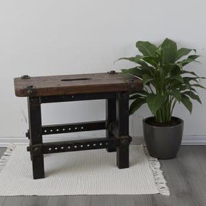 Puede incluir: Un taburete de madera rústico con una parte superior rectangular y un marco de metal oscuro. La parte superior es de madera envejecida con un acabado natural. El marco de metal tiene un diseño atornillado. Una planta en maceta está cerca.