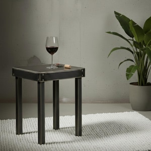 Könnte beinhalten: Kleiner, quadratischer Beistelltisch aus schwarzem Metall mit einem Glas Rotwein, einem Korkenzieher und einem Korken. Der Tisch hat vier Beine und steht auf einem weißen Teppich. Eine Topfpflanze befindet sich im Hintergrund.