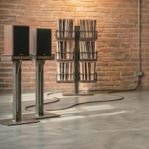 Op de afbeelding: Twee houten luidsprekers met zwarte luidsprekerfronten staan op metalen standaards. De standaards hebben een rechthoekige basis en een verticale steun. Een bakstenen muur en een metalen boekenrek vol cd's zijn op de achtergrond.