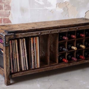 Könnte beinhalten: Ein rechteckiges Aufbewahrungsmöbel aus Holz mit einer flachen Oberseite, einem Bereich für die vertikale Aufbewahrung von Schallplatten und einem Gitterbereich für Weinflaschen. Der Rahmen besteht aus verwittertem Holz mit dunklen Metallbeschlägen.