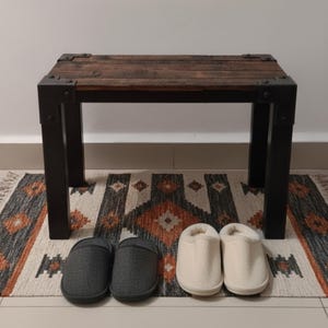 Può includere: Una piccola panca in legno con gambe e angoli in metallo scuro. Due paia di pantofole sono davanti alla panca: pantofole in tessuto grigio e pantofole bianche e soffici. La panca è su un tappeto fantasia.