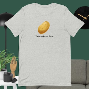 Può includere: Una t-shirt grigia melange con un'illustrazione di patata marrone e il testo "Taters Gonna Tate".