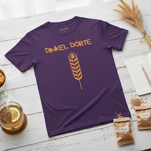 May include: A purple t-shirt with the words "DINKEL DÖRTE" in orange, and a wheat graphic. The shirt is on a white wooden table, with a cup of tea, cookies, and wheat stalks.
