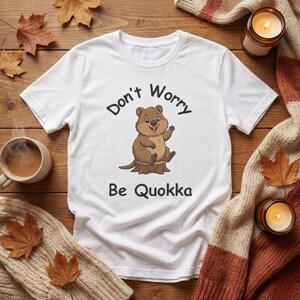 Könnte beinhalten: Weißes T-Shirt mit dem Text "Don't Worry Be Quokka" und einer Cartoon-Quokka-Illustration. Das Quokka ist braun und sitzt auf einem Baumstumpf. Das Shirt liegt auf einer Holzoberfläche mit Herbstdekorationen.