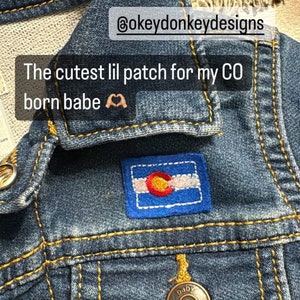 Peut inclure: Une veste en jean bleu avec un écusson rectangulaire brodé représentant le drapeau de l'État du Colorado. La veste présente des boutons en métal doré avec l'inscription baby B'gosh et des coutures contrastées jaunes sur les poches.