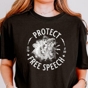 May include: Black t-shirt with a white graphic of a roaring tiger within a circle, bearing the words "PROTECT FREE SPEECH". The design also includes the text "ESTD 17 89".