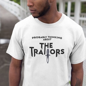 May include: White t-shirt with black text that reads "Probably thinking about The Traitors" with a black dagger graphic.